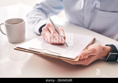 Close-up of unrecognizable businessman sitting at table with tea mug and making notes in application form Stock Photo