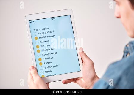 Over shoulder view of young man using tablet and checking list while packing suitcase for trip Stock Photo