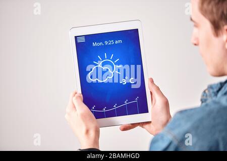Over shoulder view of young man checking weather forecast on tablet app against white wall Stock Photo