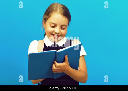 Back to school and education concept. Pupil in school uniform with smile. Girl writes in big blue book doing her homework. School girl with happy face isolated on blue background Stock Photo