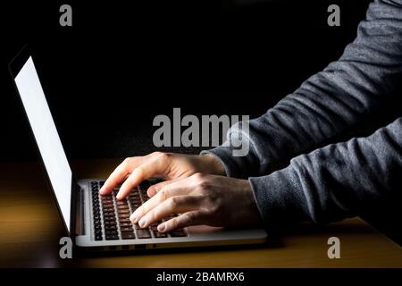 Man working on laptop with white screen glowing in the dark on a wooden desk seen from the side Stock Photo