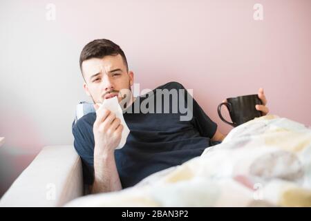 Young ill man with hanky. Guy lying on the sofa under covers. He holds napkin in one hand and black cup in other. Begins to sneeze. Isolated over Stock Photo