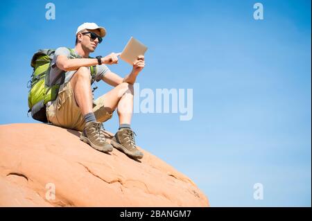 Hiker with backpack sitting outdoors using a tablet on the top of a red rock mountain Stock Photo
