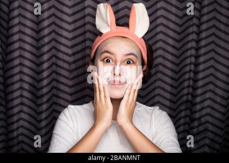 A cute and happy girl smears face with cosmetic clay or mud, standing in the bathroom, with rabbit ears on her head and smiling. The concept of health and beauty. Stock Photo