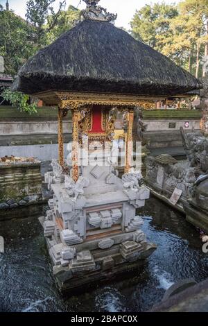 Small stone temple sitting in pool of water with golden frame, bright red clothe and thick black thatch roof in Tirta Empul Holy Water Temple Bali. Stock Photo