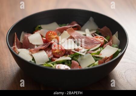 Salad with prosciutto, tomato, arugula leaves and parmesan cheese in black bowl on walnut Stock Photo