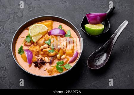 A Chicken Massaman Curry in black bowl at dark slate background. Massaman Curry is Thai Cuisine dish with chicken meat, potato, onion and many spices. Stock Photo