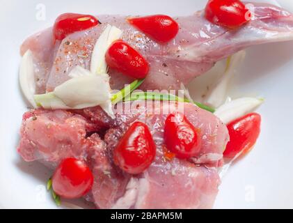 Raw Rabbit Meat, Rear Leg Cuts of Hare Seasoned with Fresh onion, cherry tomatoes and Spices in a white plate. Fresh rabbit meat Stock Photo