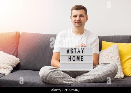 Young man sitting on the sofa in his living room holding lightbox with message Stay Home. Stock Photo