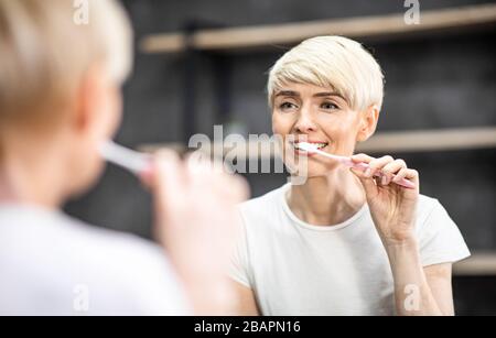 Woman Brushing Teeth Smiling To Reflection In Mirror In Bathroom Stock Photo