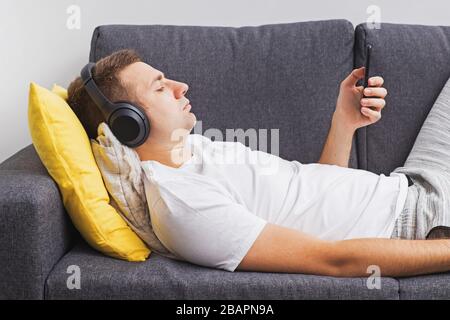 Man in wirwlwss headphones watching lying on sofa and looking on his smartphone Stock Photo