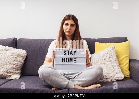Woman sitting on the sofa in her living room holding lightbox with message Stay Home Stock Photo