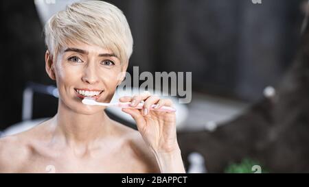 Lady Cleaning Teeth Smiling To Camera In Bathroom, Panorama Stock Photo