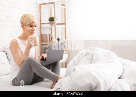 Middle-aged woman sitting in bed with laptop and cup of coffee Stock Photo