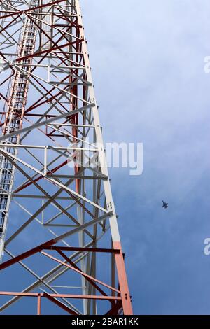 This photo shows a tall cell tower reaching up into a bright blue sky. The tower is painted in a striking red and white pattern, and you can see the d Stock Photo