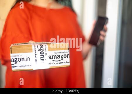 Young indian girl holding an amazon delivered box in one hand an an out of focus mobile phone in the other showing a delivery from this e-com vendor Stock Photo