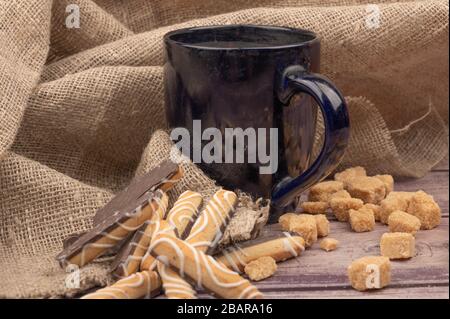 dark blue ceramic tea mug, cookie sticks with chocolate and white icing and pieces of brown cane sugar on a background of textured fabric. Close up Stock Photo