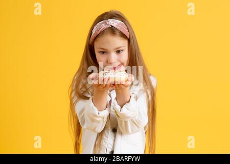 Crazy cheerful blonde girl in birthday hat smiling, having fun and looking through two red donuts on her eyes. Sweets. Yellow studio background. Stock Photo