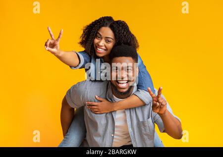 Joyful black couple embracing and showing peace sign, guy piggybacking girlfriend Stock Photo