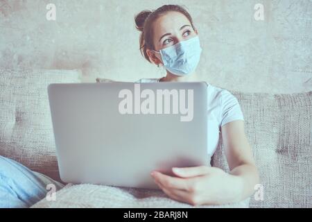 A girl in a medical mask is in quarantine at home holding a laptop and look out the window. Close up. Stock Photo