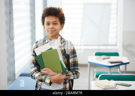 Waist up portrait of smiling African-American boy holding books and looking at camera while posing in school classroom, copy space Stock Photo