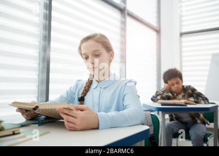 Portrait of blonde intelligent girl reading book while sitting at desk in school classroom, copy space Stock Photo