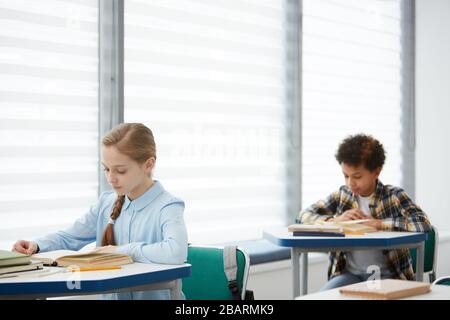 Portrait of two kids sitting in row at desks in school classroom, focus on cute girl reading book in foreground, copy space Stock Photo
