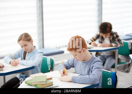 High angle view at multi-ethnic group of children sitting at desks in school and writing test notes, copy space Stock Photo