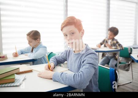 Portrait of modern red-haired schoolboy sitting at desk in classroom and looking at camera while taking notes, copy space Stock Photo
