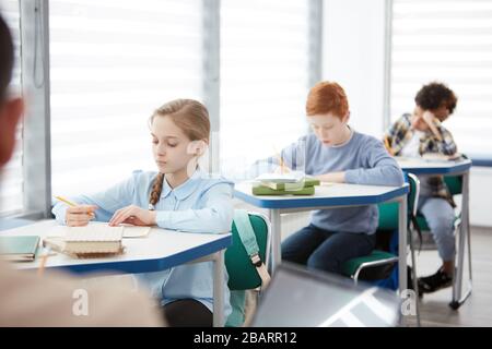 Side view at multi-ethnic group of children sitting in row at desk in school classroom and writing or taking test, copy space Stock Photo