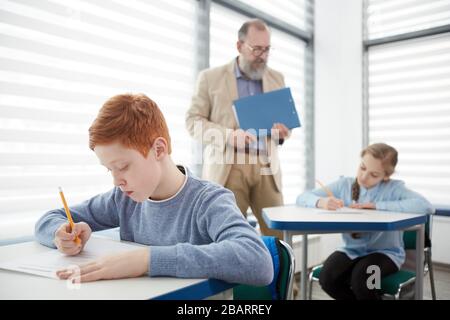 Group of children taking test with bearded mature teacher watching them, focus on red haired boy sitting at desk and writing in foreground, copy space Stock Photo