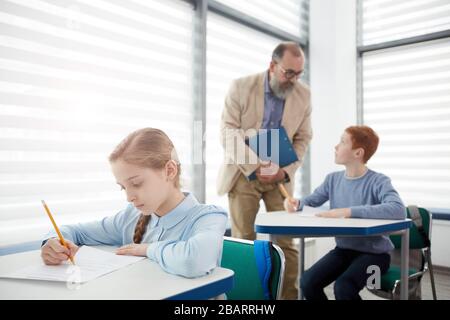 Group of children taking test with bearded mature teacher watching them, focus on cute blond girl sitting at desk and writing in foreground, copy spac Stock Photo