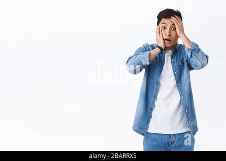 Oh my god what should I do. Shocked and concerned asian guy in panic grab his face and staring camera indecisive, standing alarmed got in terrible Stock Photo