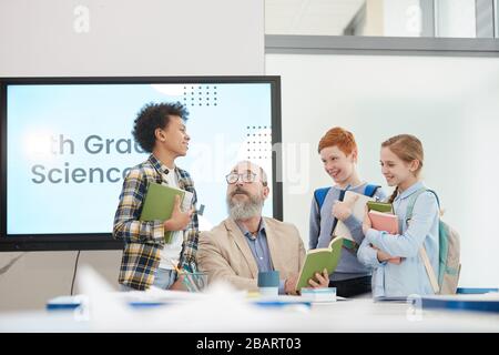 Multi-ethnic group of cheerful kids standing around bearded senior teacher in school classroom, copy space Stock Photo