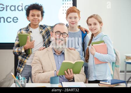 Multi-ethnic group of happy kids standing around bearded senior teacher in school classroom and looking at camera Stock Photo