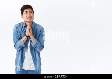 Portrait of intense young worried asian man nervously praying for dream come true, talking to God, press hands together in pray raising head in the Stock Photo