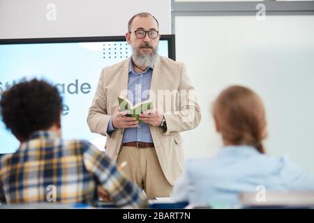 Waist up portrait of bearded senior teacher talking to group of kids in school classroom, copy space Stock Photo
