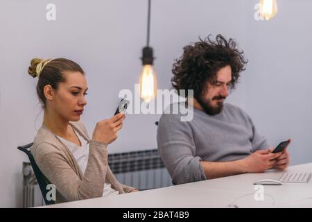 Portrait of two colleagues sitting next to each other an texting on their mobile phones inside the office Stock Photo