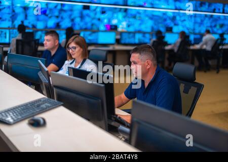 Group of Security data center operators (administrators) working in a group at a CCTV monitoring room while looking at multiple monitors ( computer sc Stock Photo