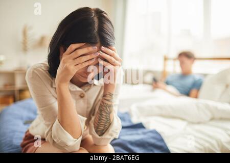 Portrait of frustrated young woman crying while sitting on bed with husband in background, couple fight, copy space