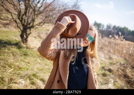 A girl in a brown coat, hat and glasses walks in a park with a lake under the bright sun. Rejoices in life and smiles. The beginning of spring Stock Photo