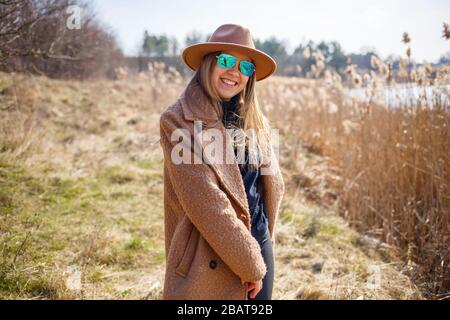 A girl in a brown coat, hat and glasses walks in a park with a lake under the bright sun. Rejoices in life and smiles. The beginning of spring Stock Photo