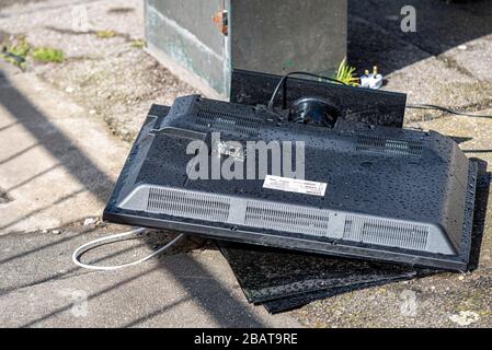 Discarded flat screen television smashed on a street pavement. Electrical waste dumped outside in the rain. Logik TV with plug attached