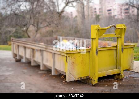 Yellow dumpster seen on a street near a park. The dumpster is filled to the top with plastic trash bags and other debris. Selective focus. Stock Photo