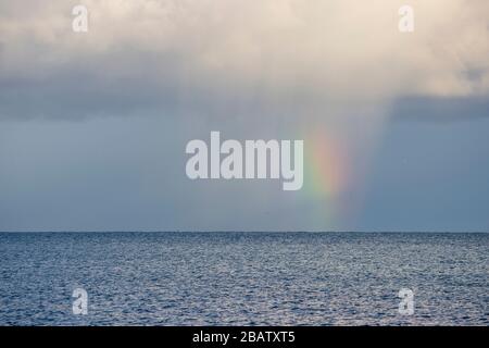 A rainbow appears as sun shines through rain clouds across the sea Stock Photo