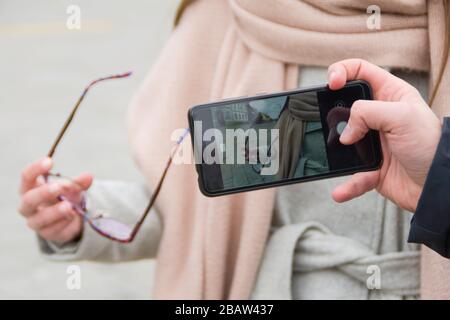 A man's hand with a smartphone is photographing the hand of a woman in a coat holding reading glasses. Stock Photo