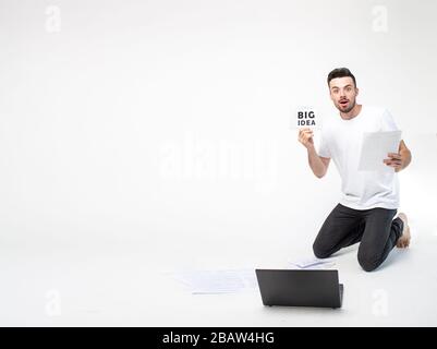 Young man isolated over background. Guy stand on knees and show big idea piece of paper. Emotional excited young man in front laptop alone in studio Stock Photo