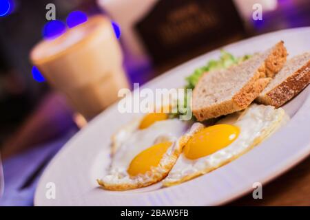 Closeup of fried eggs with toasted bread on white dish and glass with coffee on wooden table at modern restaurant. Traditional american breakfast in d Stock Photo