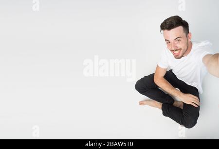 Young man isolated over background. Guy sit on floow and take selfie alone. Side view of emotional guy on picture. Stock Photo