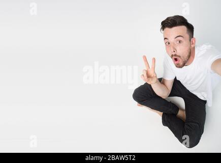 Young man isolated over background. Guy take selfie alone and sit on floor. Emotional happy with facial expression man show peace symbol. Stock Photo